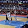 A basketball game takes place in a large indoor arena filled with spectators. Players from two teams, one in black uniforms and the other in white, compete for the ball near the hoop. The stadium is adorned with banners in Chinese, and a scoreboard displays a tied score of 21. The court features a distinctive blue and wood pattern.