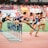A group of athletes is competing in a hurdle race on a track. Each runner is captured mid-air as they leap over barriers in a stadium setting. The background includes red stadium seating and various banners. The athletes wear numbered jerseys and running shoes.