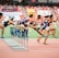 A group of athletes is competing in a hurdle race on a track. Each runner is captured mid-air as they leap over barriers in a stadium setting. The background includes red stadium seating and various banners. The athletes wear numbered jerseys and running shoes.