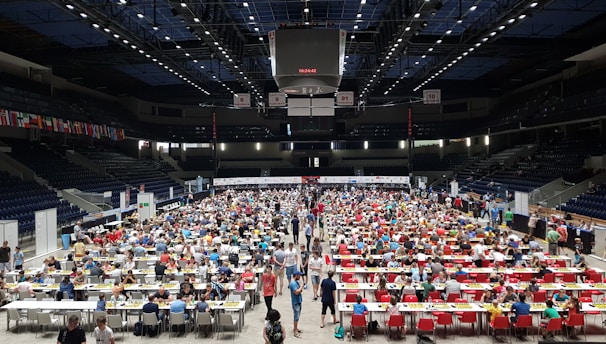 An indoor sports arena filled with rows of tables, each occupied by people engaged in competitive activities. The ceiling is high, with numerous lights illuminating the large space. The audience seating on either side is empty, and there are flags hanging from the upper level.
