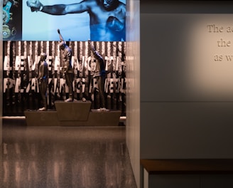 A dimly lit display features statues of athletes standing on a podium. They are positioned in front of a backdrop with the words 'LEVELING THE PLAYING FIELD' in bold letters. On the wall to the right, a softly lit quote is partially visible. The overall ambiance suggests a museum or exhibition setting.
