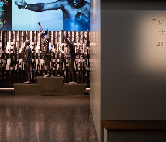 A dimly lit display features statues of athletes standing on a podium. They are positioned in front of a backdrop with the words 'LEVELING THE PLAYING FIELD' in bold letters. On the wall to the right, a softly lit quote is partially visible. The overall ambiance suggests a museum or exhibition setting.