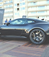 A sleek black sports car is parked on a wooden platform in an urban area. The car's design is modern and aerodynamic, with large alloy wheels and a shiny exterior. People are gathered around in the background, along with a tall banner featuring a logo. The backdrop includes a multi-story building with numerous windows.