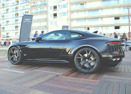 A sleek black sports car is parked on a wooden platform in an urban area. The car's design is modern and aerodynamic, with large alloy wheels and a shiny exterior. People are gathered around in the background, along with a tall banner featuring a logo. The backdrop includes a multi-story building with numerous windows.