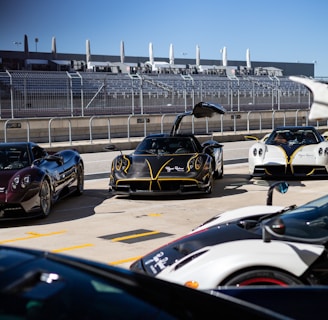 A group of high-end sports cars parked at a racetrack, with one black car prominently in front with its doors open upwards. The background features a racing facility, complete with grandstands and a metal fence. The sky is clear and blue, contributing to the overall sleek and futuristic ambiance.