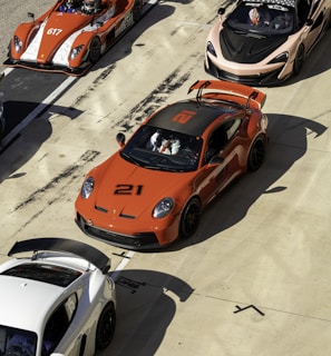 A lineup of high-performance sports cars is positioned on a racetrack. The cars are of various models and colors, including a prominent orange car with the number 21, an orange open-top car with the number 617, a sleek black car, and a white car. Drivers in racing gear are seated in some of the vehicles, preparing for a potential race. The scene conveys a sense of competition and speed, set in a sunny environment with shadows cast by the intense sunlight.