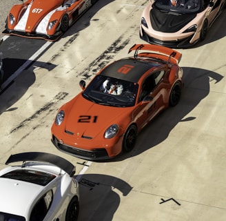 A lineup of high-performance sports cars is positioned on a racetrack. The cars are of various models and colors, including a prominent orange car with the number 21, an orange open-top car with the number 617, a sleek black car, and a white car. Drivers in racing gear are seated in some of the vehicles, preparing for a potential race. The scene conveys a sense of competition and speed, set in a sunny environment with shadows cast by the intense sunlight.