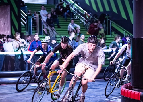 A group of cyclists competes in an indoor race, navigating a track with intense focus. They wear helmets and sport attire, with a crowd of spectators watching intently from the green bleachers in the background.