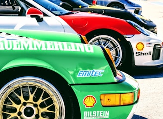 Several sports cars are lined up in a row, showcasing vibrant colors and sponsor logos. The forefront displays a green car with a detailed wheel rim and various brand decals, including Bosch and Shell. Each car features sleek designs, contributing to a dynamic and competitive atmosphere.