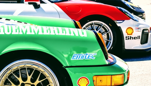 Several sports cars are lined up in a row, showcasing vibrant colors and sponsor logos. The forefront displays a green car with a detailed wheel rim and various brand decals, including Bosch and Shell. Each car features sleek designs, contributing to a dynamic and competitive atmosphere.