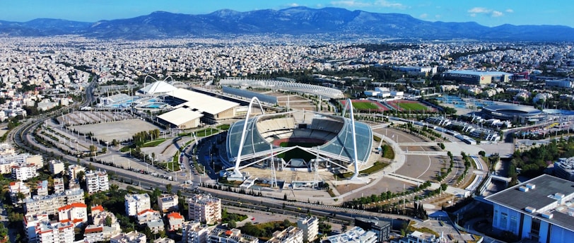 A large, modern sports complex is situated within an expansive urban landscape. The main stadium features prominent architectural structures and an open roof design. Surrounding the stadium are various sports facilities, roads, and greenery. Residential and commercial buildings extend into the distance, framed by mountain ranges under a clear blue sky.