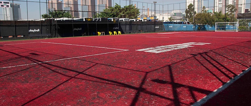 A sports court with a red playing surface and white lines, surrounded by a black mesh fence. In the background, tall buildings are visible, along with some trees and a few yellow chairs positioned near the fence.