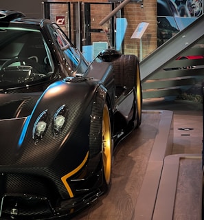 A sleek and luxurious sports car with a carbon fiber body and gold alloy wheels is displayed on a polished wooden platform. The setting includes glass barriers, informational plaques, and a staircase in the background, creating a sophisticated showroom environment.