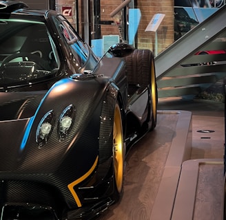 A sleek and luxurious sports car with a carbon fiber body and gold alloy wheels is displayed on a polished wooden platform. The setting includes glass barriers, informational plaques, and a staircase in the background, creating a sophisticated showroom environment.